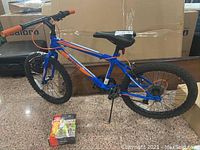 Full view of the blue and orange children's bicycle with 20-inch tires and orange grips, with boxed training wheels on the floor in front