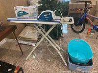 Ironing board with iron on top, blue and gray plastic baskets on the floor next to it, other items seen on sides.