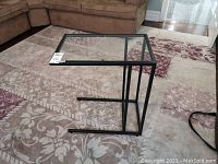 Portable laptop table with black metal frame and glass top, positioned on a carpet with a sofa nearby.
