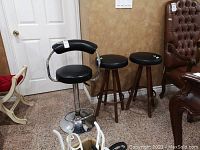 Photo showing two wooden stools with black faux vinyl padded seats alongside one metal bar stool with black padded seat and backrest with armrests, chrome finish.