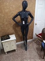 Full body image of shiny black female mannequin standing with hands on hips against a wall.