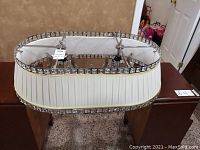 Oval chandelier resting on a wooden table with a white pleated fabric shade bordered with glass and chrome square elements and crystal dangles hanging inside.