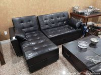 Full view of two-piece black faux leather sectional couch arranged as a sectional in a room with tile flooring and decorative items nearby.