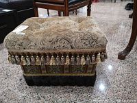 Front top view of fabric square ottoman with paisley and floral pattern fabric, with ornamental tassels along edge and black faux fur bottom section.