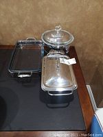 Three chafing dishes set on a table: two rectangular glass dishes with stainless steel frames and lids, one round stainless steel dish with lid and stand.