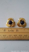 Pair of gold-toned metal earrings with deep purple center stones, measured against a ruler showing approximately 3/4 inch size.