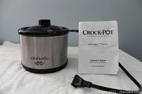 Small Crock-Pot Little Dipper Warmer with stainless steel body, black lid, powered cord and owner's guide booklet placed on a white cloth surface.