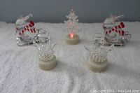 Photo shows five Christmas ornaments arranged on a white towel: two glass snowmen with bead details on white bases, two glass Santas in red and white striped sleighs, and one small lit plastic Christmas tree emitting red light.