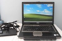 Front view of Dell Latitude D620 laptop powered on showing Windows XP desktop with power adapter and cord beside it.