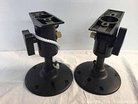 Two black 3-axis wall mounts for speakers or cameras shown from front angle. One mount has a white security cable attached.