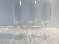 Three clear crystal champagne flute glasses standing upright on a light-colored cloth, showing their elongated bowl shape and thin stems.