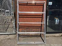 Full view of the welded aluminum shelving unit set upright outdoors against a brick wall and a chain link fence.