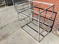 Welded aluminum square tube shelves frame photographed outdoors against a brick wall and chain link fence, showing side and front views.