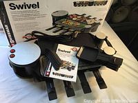 Overall view of Swissmar Swivel raclette party grill components including grill plates, base unit with power cord, 8 raclette pans, and instruction manual, with the product box in background.
