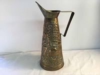 Side view of vintage brass carafe pitcher showing embossed floral and architectural relief designs and handle shape.