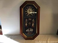 Front full view of the octagonal wooden framed nautical wall clock showing clock face and rope knot decorations, as well as miniature ship model below.