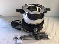 Electric fondue set showing stainless steel fondue pot seated on black heating base with attached power cord and temperature control dial, 8 fondue forks bundled in plastic packaging in front.