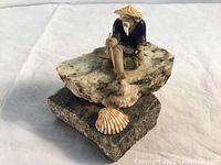 Figurine of an old Asian man sitting on two granite stones with a seashell in front, viewed from the front and slightly angled.