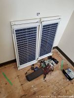 Two framed solar panels leaned against wall with controller, small panel and cables on floor