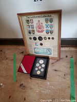 Full view of framed Centennial Collection including coats of arms, coins, a one dollar bill, and stamps. Royal Canadian Mint coin set case visible on floor.
