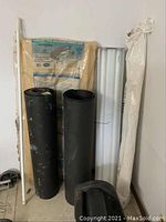 Tar paper rolls, fluorescent light fixture, vapor barrier plastic rolled, and package of ventilation channels leaning against wall on floor.