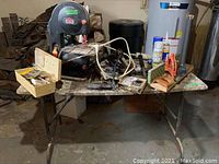 Wide view of the folding table with Jobmate band saw and assorted tools and hardware arranged on top. Includes router cutter case, hacksaws, hand tools, canisters, and various accessories.