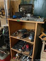 Wooden cupboard with two shelves containing various power tools and accessories; tools on top of cupboard clearly visible; mixture of cords and hand tools seen