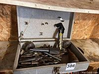 Open metal case containing multiple large drill bits and tools, placed on a wooden surface.