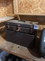 Closed black metal toolbox on wooden shelf showing wear, latches, and handle.