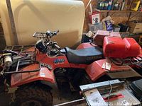 Full side-angle view of red Suzuki QuadRunner ATV in a garage, rear rack loaded with two red marine-style gas cans.