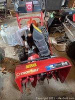 Front view of snowblower showing red housing, auger assembly, tire chains, and gas can in background