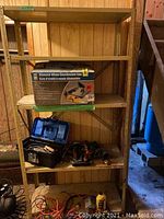 Full view of metal shelving unit showing boxed Diamond Wheel Benchmont Saw, black toolbox with hand tools, and power tools on shelves in a wood-paneled room.