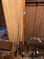 Photo showing five fishing rods standing against a wood-paneled wall, reels attached to rods visible at handles, some handles cushioned with black grips.