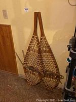 Pair of traditional wood frame snowshoes with nylon strap webbing and bindings leaning against a wall, showing overall condition and size.