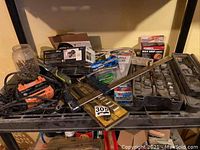 Full view of all items on black metal shelf including battery charger, jar of nails, clamp, socket set, packaged night light, extension cord.