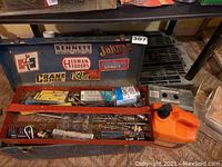 Front view of open vintage green metal toolbox with automotive-themed stickers on lid. Inside contains drill bits, a can of pipe cement, and other tools in compartments.