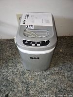 Front view of the silver RCA ice maker with control panel and transparent lid, user manual on top.