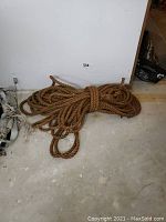 Long, thick twisted natural jute rope coiled on concrete floor against white wall, showing size and texture; rope looks heavy-duty with natural fibre composition