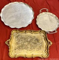 All three trays displayed together on a red cloth: two hammered aluminum trays, one large round and one smaller with handles, and one ornate gold-toned rectangular tray.