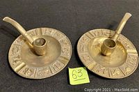 Pair of vintage brass ashtrays with pipe figures and zodiac engravings on edges, shown from top angle.