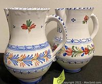 Photo showing two hand-painted ceramic pitchers with blue trim, floral designs, bulbous bodies, and handles.
