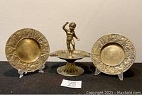 Full shot showing two round zodiac brass ashtrays flanking footed brass ashtray with figurine.
