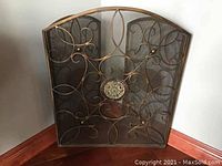 Front view of brass fireplace screen showing curved top, ornate scrollwork, and central medallion.