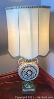 Front view of the vintage porcelain and brass table lamp with cream scalloped fabric lampshade lit.