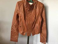 Brown genuine leather ladies jacket hanging on a green hanger, showing front lace-up detail and snap button pockets.