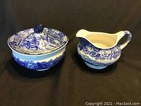 Lidded sugar bowl and creamer shown side by side against black background highlighting blue and white floral and village scene design.