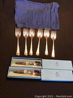 Six silver-toned dessert forks with four tines arranged in a row with a cloth and two boxed Birks tea spoons below them.