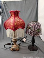 Two desk lamps on draped cloth background; one with red and gold Art Nouveau style fabric shade with fringe, and one with pink stained glass rose pattern shade on dark metal base.