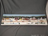 Full view of the 6x24 inch folk art acrylic painting depicting a winter village scene with buildings, cars, and people.