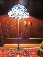 Full view of lamp showing metal base, pole and shade lit against wood door and rug